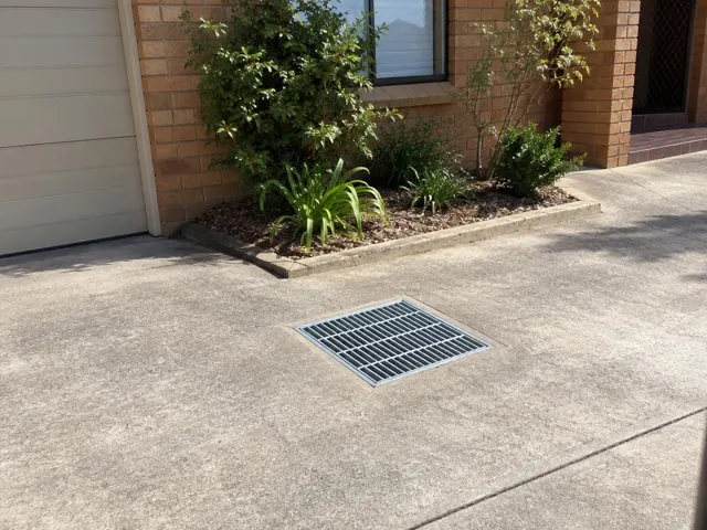Outdoor driveway drain grate beside a brick house with dry concrete paving in Kingsgrove